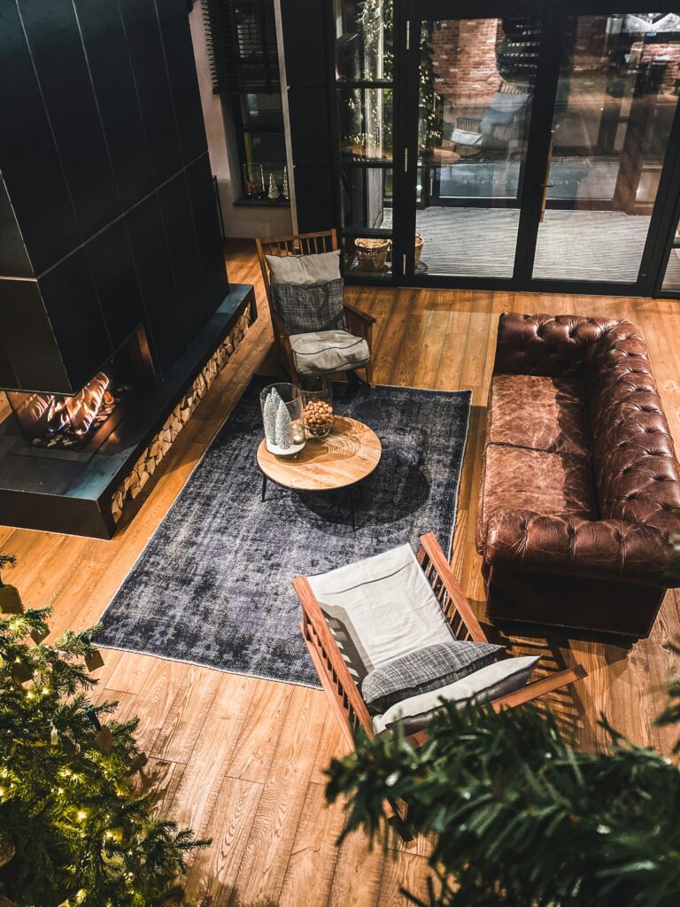 Cozy living room with a brown leather sofa, wooden chairs, a small round coffee table, and an indoor plant. The room features a dark rug on light wooden flooring and large glass doors in the background.