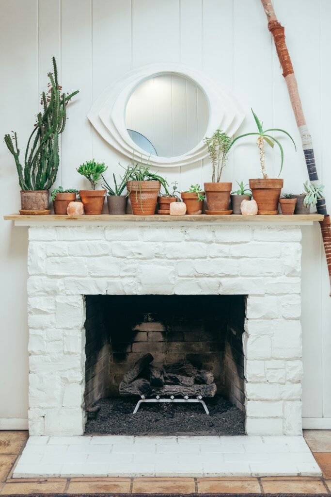 White-painted brick fireplace with various potted plants on the mantelpiece, a round mirror above, and decorative logs inside the fireplace