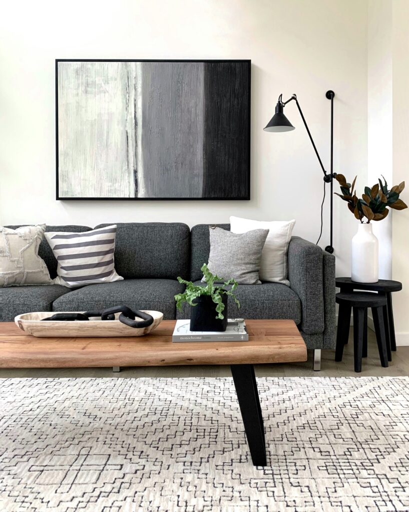 A serene and stylish living space featuring a gray sofa with various cushions, a large abstract painting above, a wooden coffee table with decor, and a black floor lamp. The room is enhanced with plants and a geometric-patterned rug.